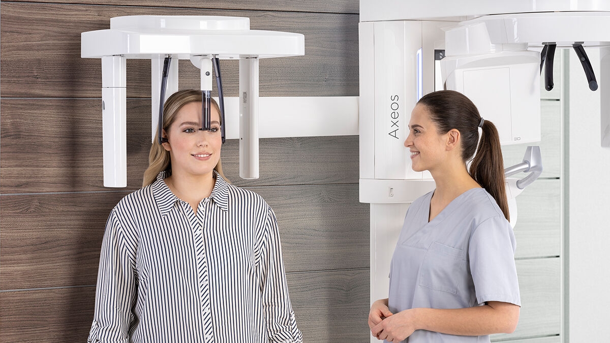 Woman standing under the cephalometric arm of Axeos and dental assistant next to her